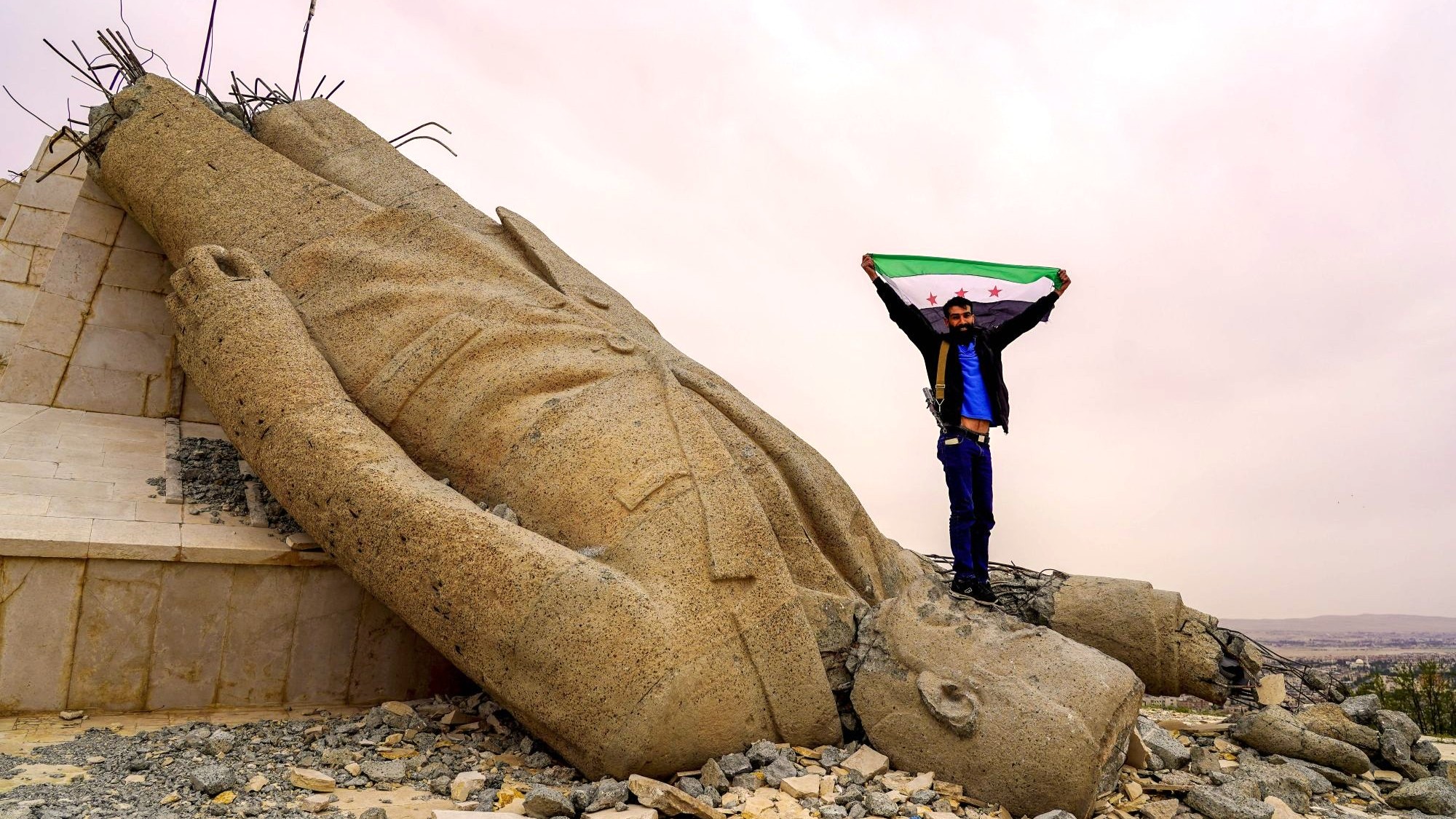 دير عطية في منطقة دمشق، يناير/كانون الثاني 2025. (Picture Alliance / SIPA | L. Barakat) رجل يرفع علم سوريا الحرة واقفًا على رأس تمثال أسد مقلوب.