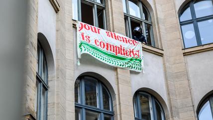 Die Fassade eines Gebäudes mit einem Schild an den Fenstern, auf dem steht: "Euer Schweigen bedeutet Mitschuld".